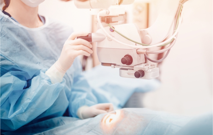 A female ophthalmologist positions a LASIK excimer laser over the eye of a patient fully draped in a surgical gown.