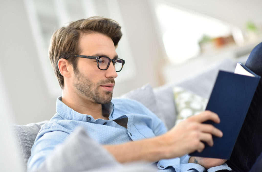 Young man with glasses reading a book on a couch.