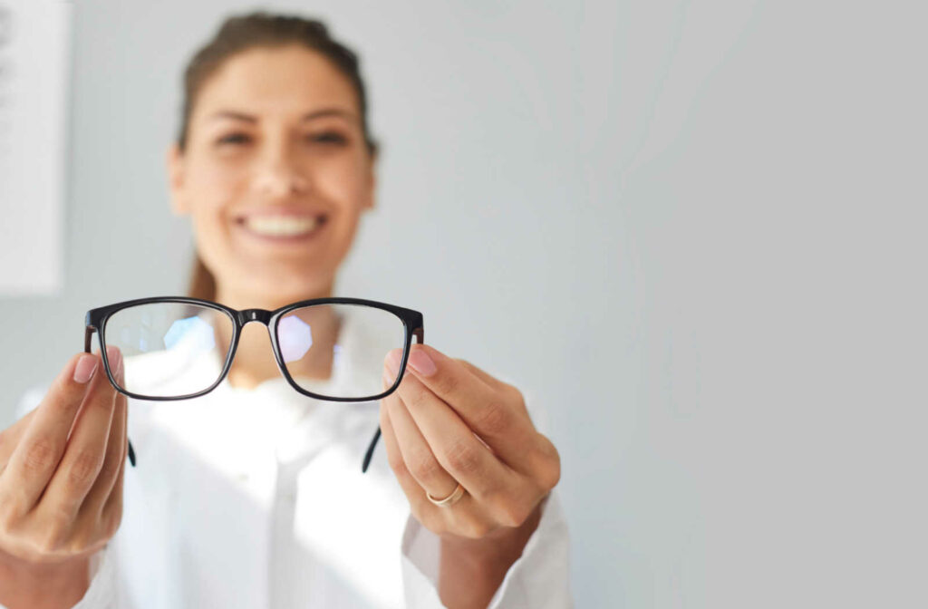 An optometrist holding a pair of prescription glasses as she smiles and looks directly at the camera.