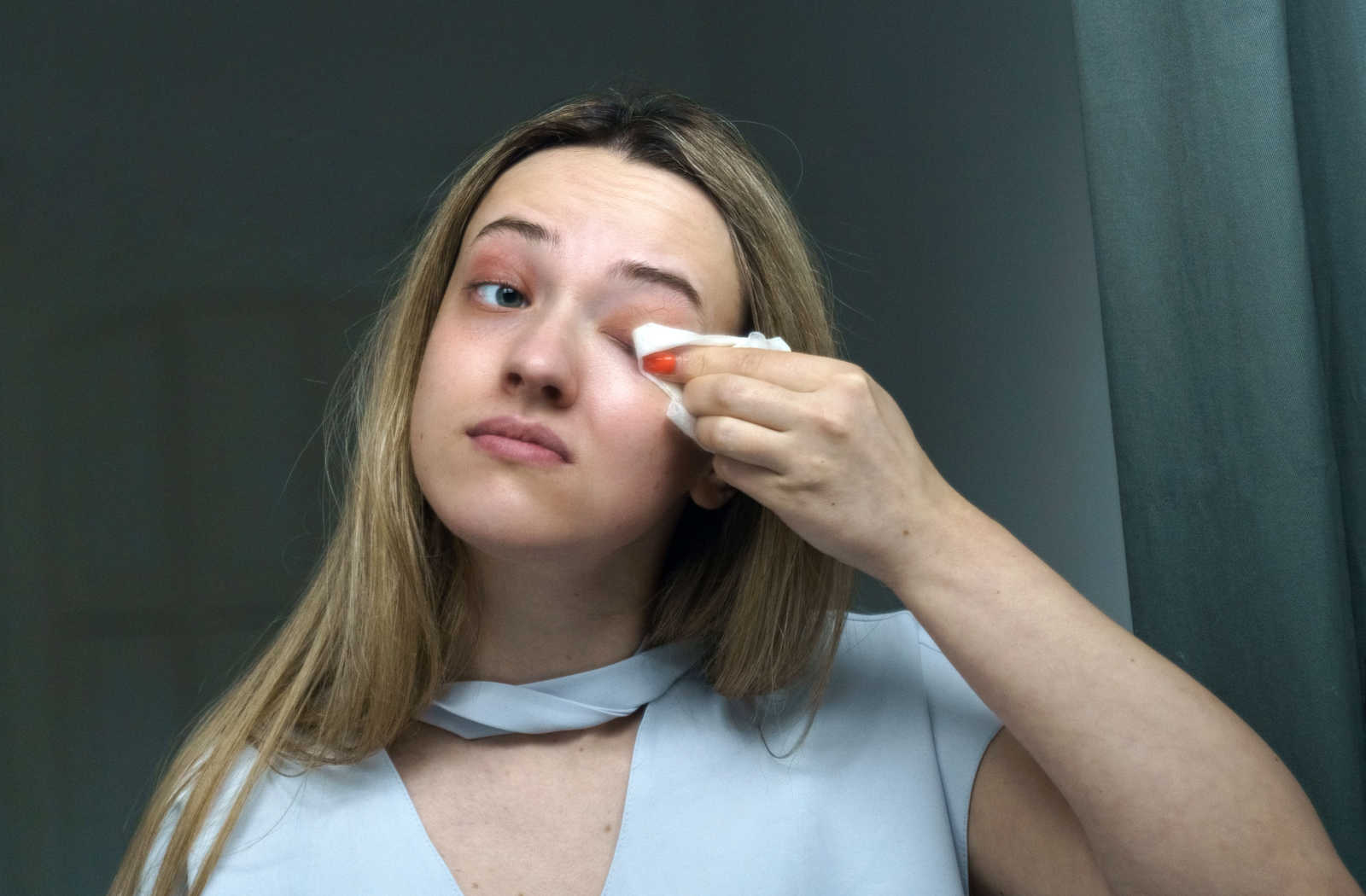 A young blonde woman cleaning her eyelids with a clean wipes using her left hand.