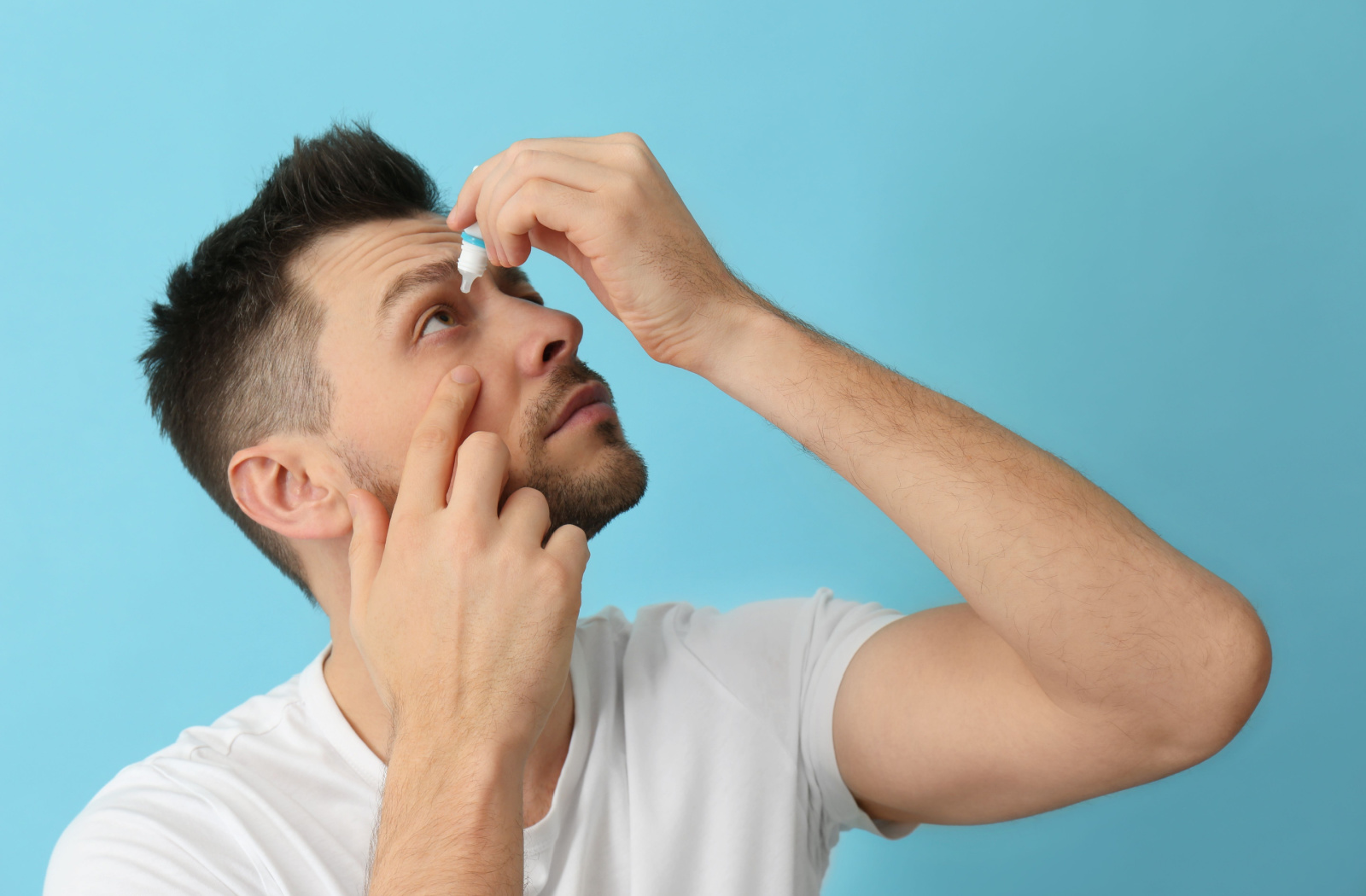 A man holding a small bottle of eye drops in his left hand and putting them on his right eye while he uses his right index finger to pull his eyelid down.