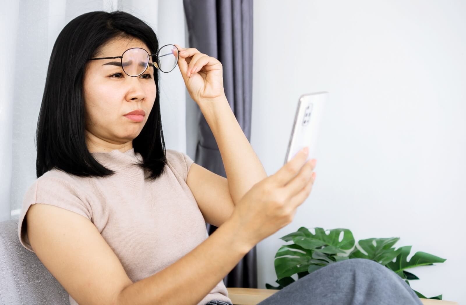 Asian woman struggles to see her phone screen, so she removes her glasses.