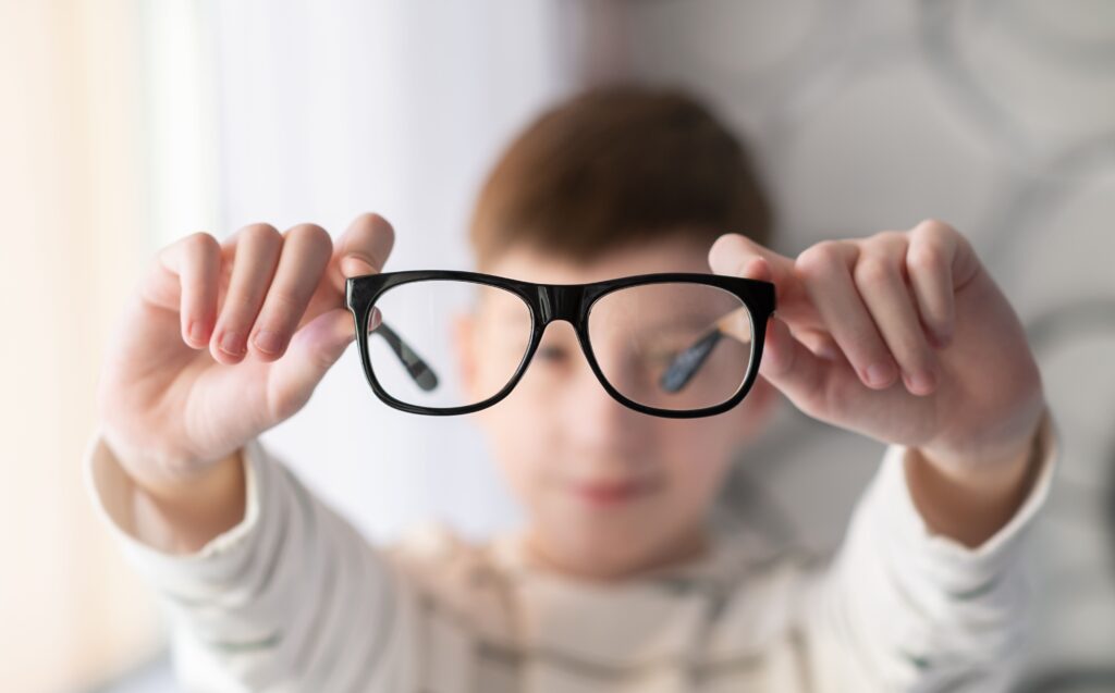 A child holding up a pair of black-rimmed eyeglasses in front of their face, with the glasses in sharp focus and the child blurred in the background.