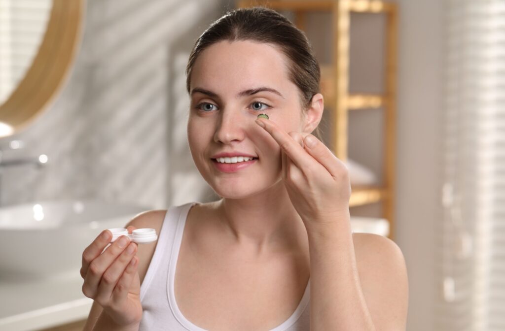 A young woman holds a contact lens case in one hand while applying a contact lens to her eye with the other hand.