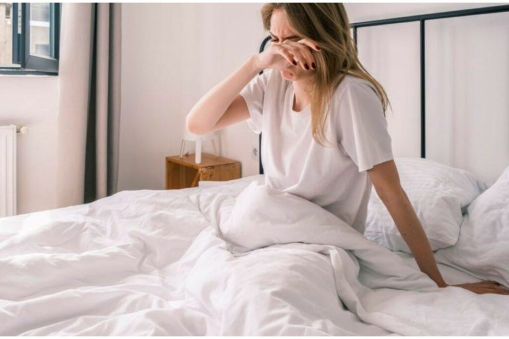 A young woman sitting up in a white bed in a brightly lit room, looking distressed and rubbing her eye with her hand, suggesting discomfort, dryness, or allergy symptoms, as seen in a medical or eye care context.